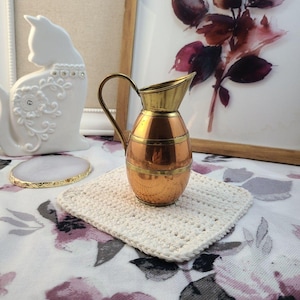May include: A small copper and brass pitcher with a handle sits on a square, cream-colored crocheted coaster. The pitcher has a rounded body with horizontal brass bands. A white ceramic cat figurine and a framed floral print are in the background.