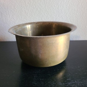 Può includere: Una ciotola rotonda in ottone metallico con una finitura liscia e lucida e un bordo largo leggermente svasato. La ciotola presenta un interno profondo e aperto e una base arrotondata, mostrando una calda tonalità giallo oro con sottili variazioni.