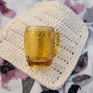 May include: A small, amber-colored glass mug with a handle, resting on a cream-colored, crocheted coaster. The mug has a textured, barrel-like design. The coaster is square-shaped with a textured pattern. The background features a floral print.