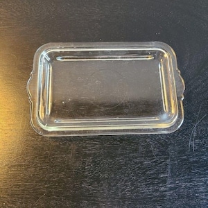 May include: A clear, rectangular glass butter dish with a lid. The dish has a slightly raised rim and decorative handles on each end. The glass is transparent, allowing the contents to be visible. The dish is designed to hold butter or other similar items.