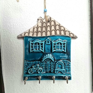 Puede incluir: Un llavero de cerámica turquesa con forma de casa con un techo texturizado y un diseño de bicicleta. El llavero tiene tres ganchos de metal y cuelga de una cuerda con cuentas. La casa presenta ventanas, una puerta y un diseño arqueado.