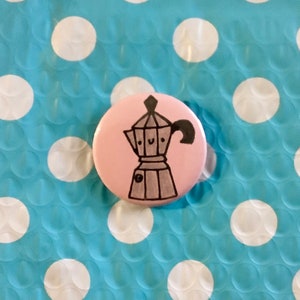 May include: A pink button with a black and white illustration of a coffee pot with a smiling face.
