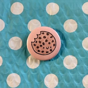 May include: A pink button with a brown chocolate chip cookie drawn on it. The cookie has a bite taken out of it and a smiley face drawn on it. The button is on a blue background with white polka dots.