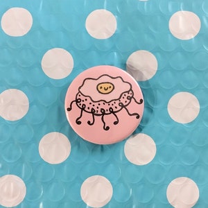 May include: Pink button with a white and yellow cartoon fried egg with a smiling face and black legs.