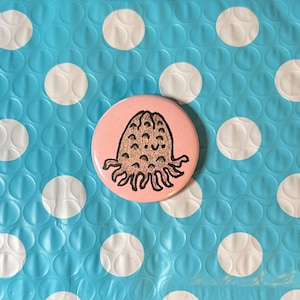 May include: A round, pink button featuring a black and brown cartoon octopus design. The button is set against a blue background with white polka dots, creating a playful contrast. The button is a fun accessory.