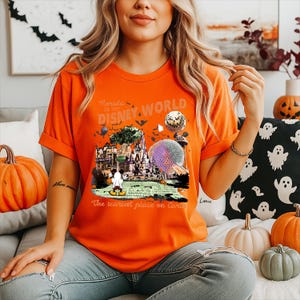 Puede incluir: Una camiseta naranja de manga corta con un estampado gráfico de monumentos de Disney World, un personaje fantasma y murciélagos. El texto dice Florida Est 1971 Disney World y The scariest place on Earth.