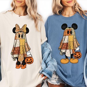 以下が含まれることがあります： クリーム色のTシャツと青いTシャツ。どちらもパッチワーク柄のシーツを被って幽霊に扮した漫画キャラクターのグラフィックが描かれています。キャラクターはハロウィンのキャンディを入れるためのオレンジ色のカボチャのバケツを持っています。