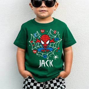 Op de afbeelding: Een groen t-shirt met een Spider-Man-ontwerp. Het ontwerp toont een rood en blauw Spider-Man-personage in een web, met harten, sneeuwvlokken en de naam "JACK" in het wit. Het kind draagt een zwarte zonnebril en een geruite korte broek.