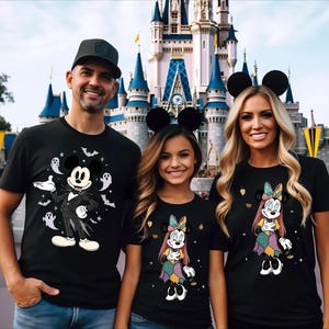 Könnte beinhalten: Drei schwarze T-Shirts mit Grafikdrucken von Figuren in Nadelstreifenanzügen und Patchwork-Kleidern, umgeben von Geistern, Fledermäusen und Sternen. Die T-Shirts werden mit blauen Jeans und schwarzen Mausohr-Haarreifen vor einem Schloss getragen.