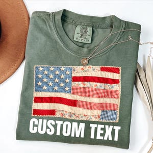 May include: An olive green t-shirt with a patchwork American flag design. The flag features blue, red, and white fabric with stars and stripes. The text "CUSTOM TEXT" is printed in white below the flag. A silver necklace with a pendant is draped on the shirt.