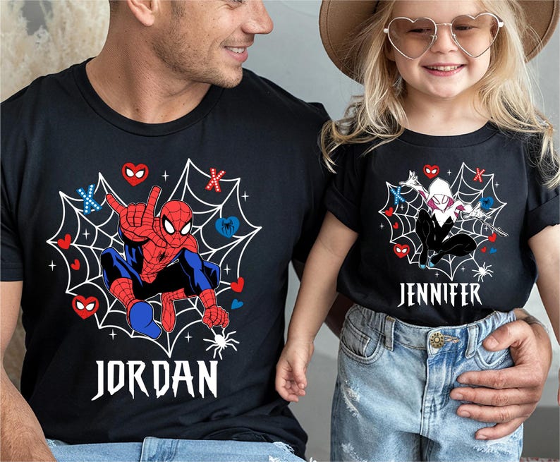 Op de afbeelding: Zwarte T-shirts met Spider-Man en Spider-Gwen ontwerpen. Het Spider-Man shirt heeft de naam "JORDAN" in het wit, terwijl het Spider-Gwen shirt "JENNIFER" heeft. Beide shirts hebben een webdesign met harten en spinnen.