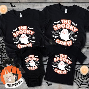 Puede incluir: Una colección de camisetas negras y un body de bebé con un gráfico de fantasma blanco, telarañas y murciélagos. El texto dice THE SPOOKY CREW en letras naranjas y blancas. Se incluye una pequeña bolsa de tela con el mismo diseño.