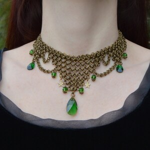 May include: A bronze-toned chainmail necklace with green gemstone accents. The necklace features a woven chain design, teardrop-shaped green gems, small star charms, and smaller green beads. The necklace is worn against a black background.