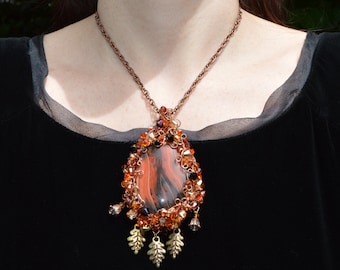 May include: A copper necklace with a large, oval-shaped pendant featuring a brown and black agate stone. The pendant is surrounded by copper wire and small, amber-coloured beads. The necklace is accented with small, gold-coloured leaves.
