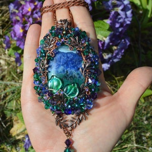 Purple flower pendant, wire wrapped in copper and tiny blue, purple and green crystals. Central agate stone is marbled rich blue purple, with dark greens across bottom half. 2 glass leaves, and a mass of tiny beads are woven into the wire wrap.