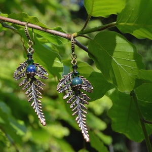 Puede incluir: Un par de pendientes de helecho de cobre con detalles de piedras preciosas verdes y azules. Los pendientes est&aacute;n colgados de una rama.