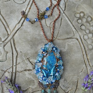 Blue crystal wirewrap pendant laid on a table to show the long chain. Pendant is made of copper, with a richly textured enctral stone in blue an white. Beads, crystals and tiny flowers surround the stone, and dangle underneath.