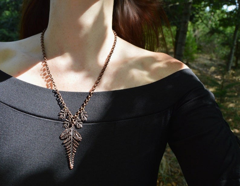Puede incluir: Un collar de cobre con un colgante de helecho. El collar lo lleva un maniqu&iacute; con un top negro de hombros descubiertos.