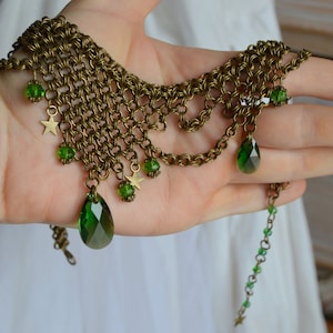 May include: An antique brass-toned necklace with a chainmail design. The necklace features green teardrop-shaped and round gemstones, along with small star charms. The necklace is displayed on a hand.