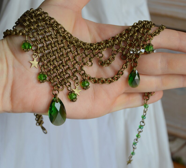 May include: An antique brass-toned necklace with a chainmail design. The necklace features green teardrop-shaped and round gemstones, along with small star charms. The necklace is displayed on a hand.