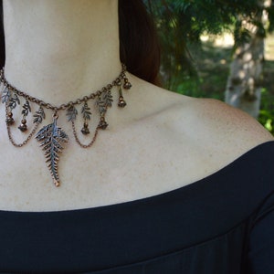 Beaded choker with fern leaves and large copper fern in the middle, on a thick copper chain. Smaller loops of chain and beads hang from the little ferns either side. The wearer in a black dress is standing in the shade of trees.