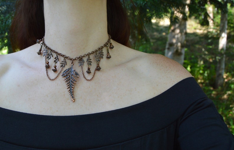 Beaded choker with fern leaves and large copper fern in the middle, on a thick copper chain. Smaller loops of chain and beads hang from the little ferns either side. The wearer in a black dress is standing in the shade of trees.