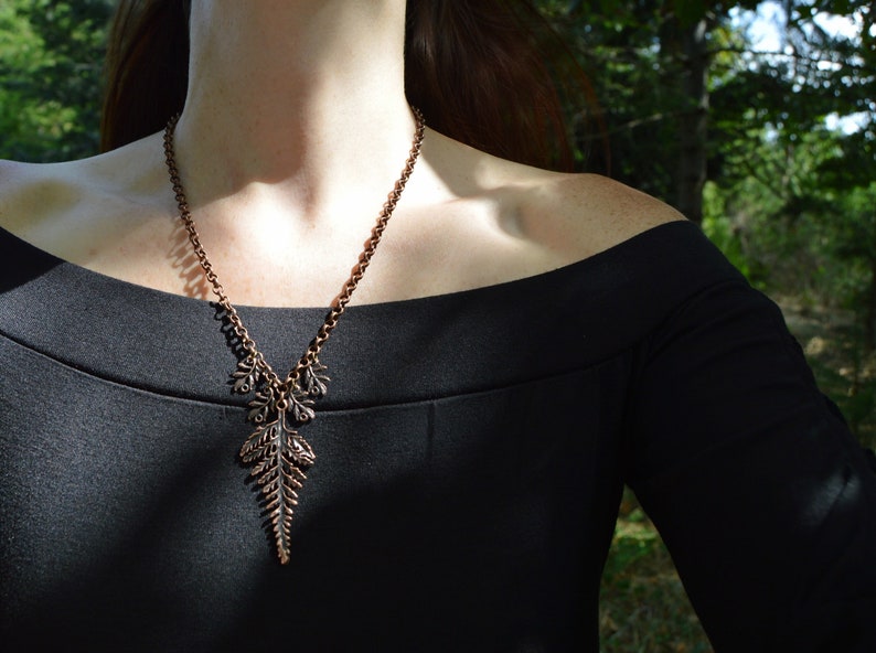 Copper leaf pendant with fern on a long chain, shown worn across the neck / chest of a person in a black dress standing in a forest. The central fern leaf is large at 2 1/2 inches long. abive it hang 4 small fern leaves, 2 on each side.