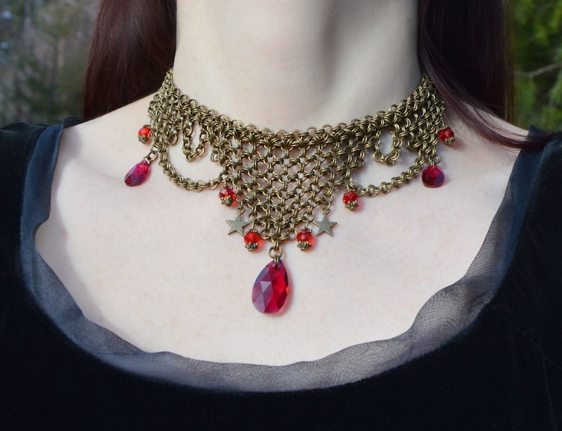 May include: A brass-toned choker necklace with a chainmail design. The necklace features dangling red teardrop-shaped gems, small red beads, and star-shaped charms. The necklace is worn against a dark background.