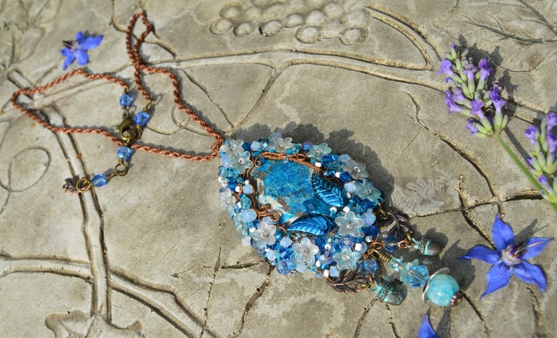 Blue and white flower necklace made of copper wire, little crystals and beads. The central stone is medium blue and white with a marbled texture. Little white flowers, crystals and blue leaves surround it. Beaded pendants hang underneath.