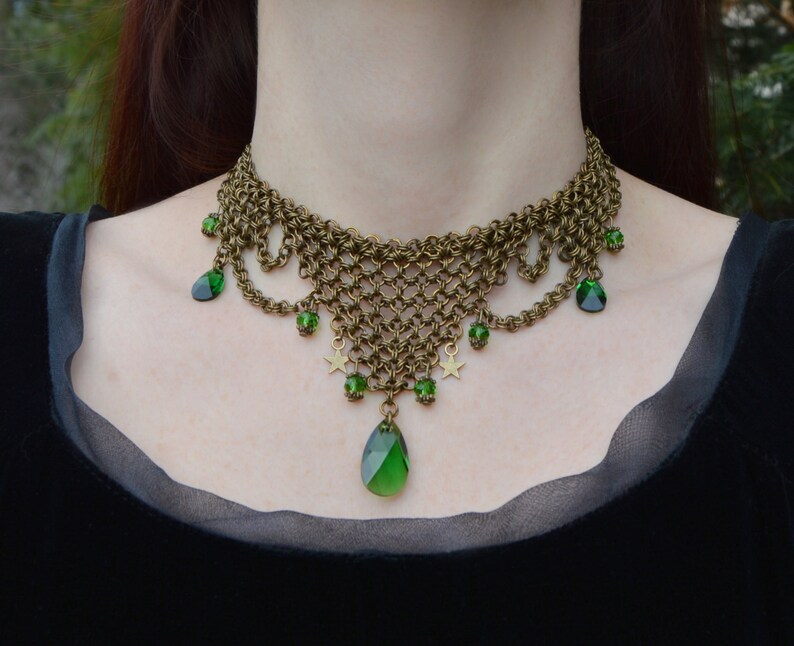 May include: A bronze-toned chainmail necklace with green gemstone accents. The necklace features a woven chain design, teardrop-shaped green gems, small star charms, and smaller green beads. The necklace is worn against a black background.