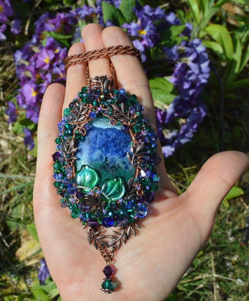 Purple flower pendant, wire wrapped in copper and tiny blue, purple and green crystals. Central agate stone is marbled rich blue purple, with dark greens across bottom half. 2 glass leaves, and a mass of tiny beads are woven into the wire wrap.