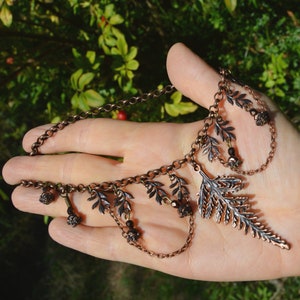 Copper fern choker necklace, displayed on a hand for size. The large central fern leaf is 2 1/2 inches long, it hangs from a copper chain with tiny fern leaves dotter either side. Beads and loops of chain hang down from these small ferns.