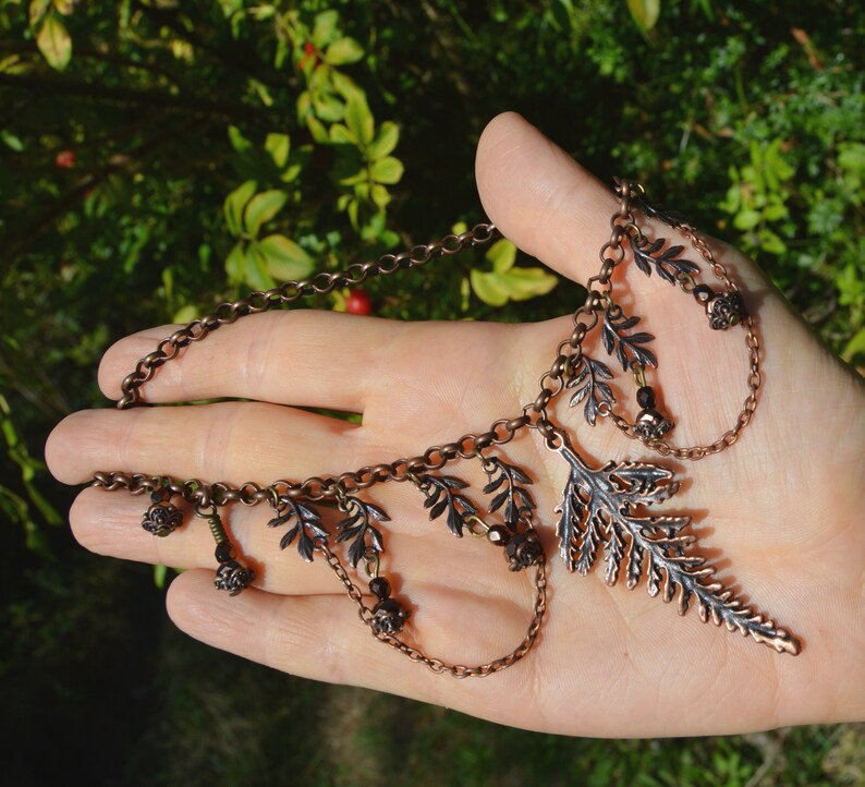 Copper fern choker necklace, displayed on a hand for size. The large central fern leaf is 2 1/2 inches long, it hangs from a copper chain with tiny fern leaves dotter either side. Beads and loops of chain hang down from these small ferns.