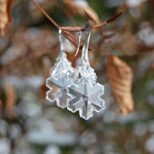 large crystal snowflake earrings, made of clear glass beads in a snowflake shape, with a frost rime of smaller crystals hanging from silver plated hooks. Earrings are shown against a winter scene, refracting the light to show the 3D snowflake shape