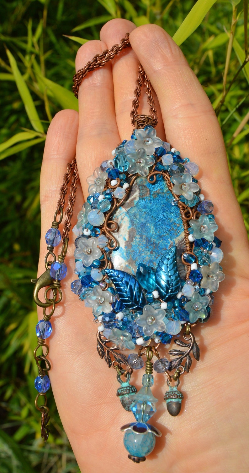 Blue and white beaded wire wrap pendant shown on a hand. The pendant from top to bottom nearly covers the hand. A range of milky whites and light blue crystals dot the pendant, along with small white flower beads.