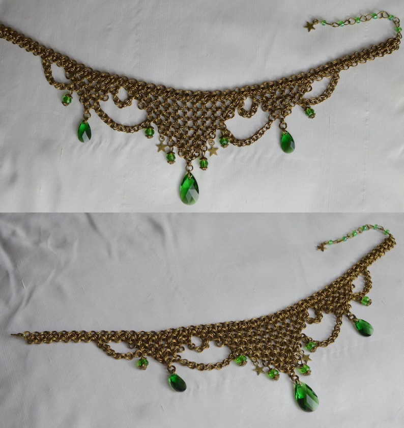 May include: A gold-toned chainmail necklace with green teardrop-shaped and star-shaped accents. The necklace features a delicate, lacy design with multiple green gems hanging from the bottom edge. The chain extends to a clasp with additional green beads and a star.