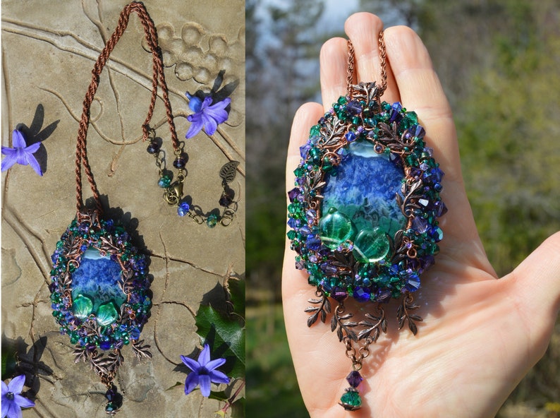 Purple and green fairy pendant, shown laid flat and in a hand for scale. Pendant is large at 4 and half inches long, oval in shape and elaborately wire wrapped in copper, tiny crystals and leaves. smaller leaves dangle underneath.