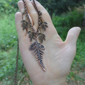 Puede incluir: Un collar de cobre con un colgante de helecho y peque&ntilde;os abalorios de hojas. El collar est&aacute; en una cadena con un abalorio de estrella en la parte superior.