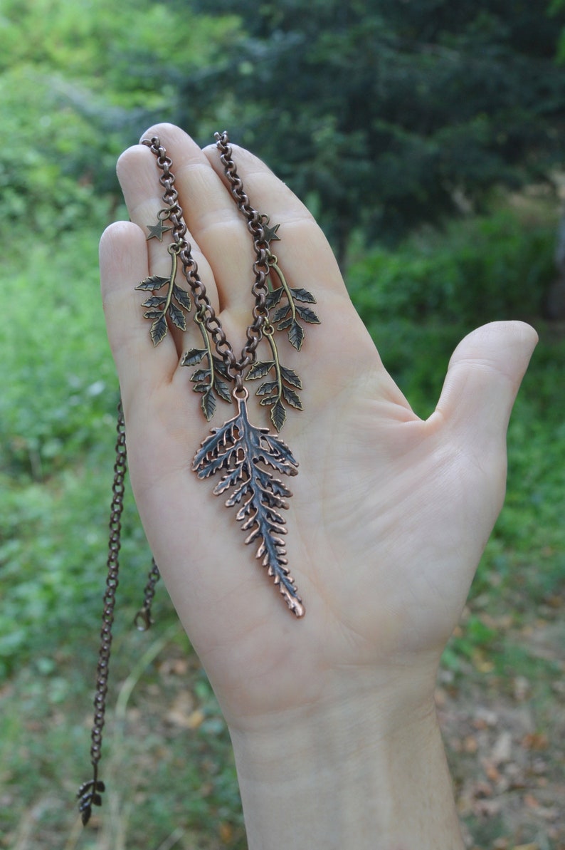 Puede incluir: Un collar de cobre con un colgante de helecho y peque&ntilde;os abalorios de hojas. El collar est&aacute; en una cadena con un abalorio de estrella en la parte superior.