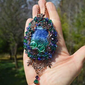 Green purple flower necklace, on a hand in natural detail. A mass of tiny crystals are woven into the wire wrap, and glitter faintly in the light. The central agate is marbles blue and green, rich colours reflected in the beadwork.