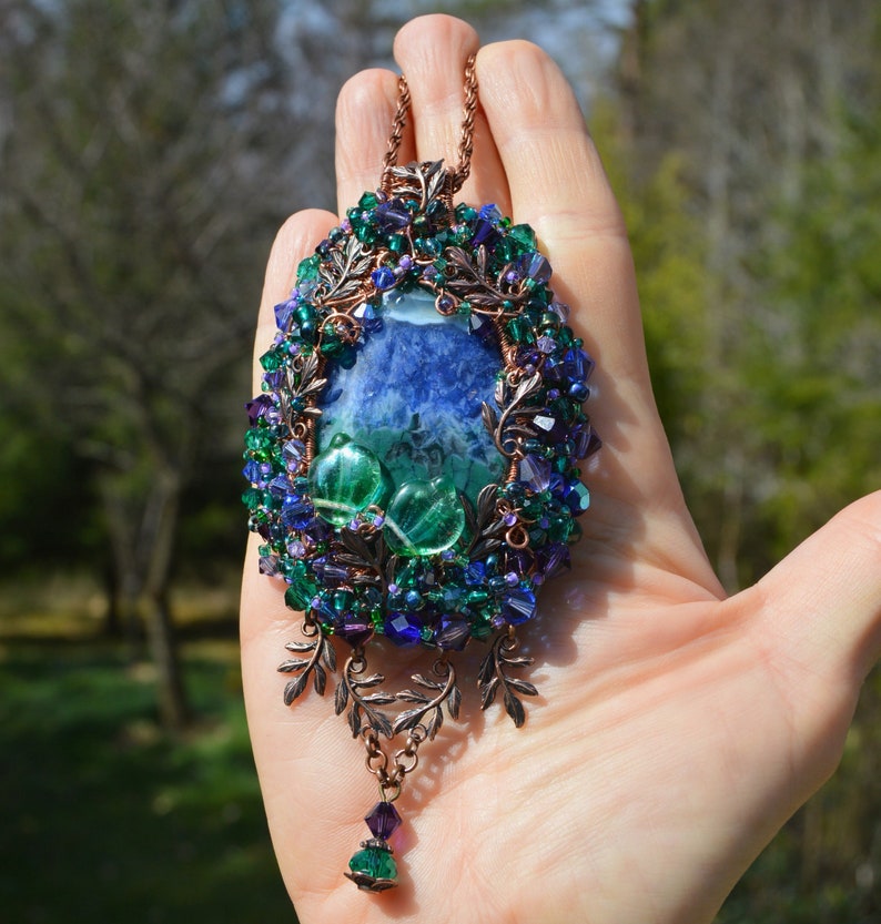 Green purple flower necklace, on a hand in natural detail. A mass of tiny crystals are woven into the wire wrap, and glitter faintly in the light. The central agate is marbles blue and green, rich colours reflected in the beadwork.