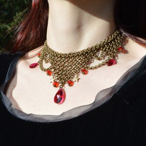 May include: A bronze-toned choker necklace with a woven chain design. The necklace features dangling red teardrop-shaped gems and smaller red beads. Small gold star-shaped charms are also incorporated into the design. The necklace is worn against a black garment.