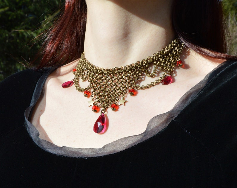 May include: A bronze-toned choker necklace with a woven chain design. The necklace features dangling red teardrop-shaped gems and smaller red beads. Small gold star-shaped charms are also incorporated into the design. The necklace is worn against a black garment.