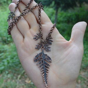 Copper leaf pendant displayed on a hand. Large central fern leaf is 2 1/2 inches long, with smaller ferns either side hanging from a thick copper chain. Behind is lush green of a summer forest.