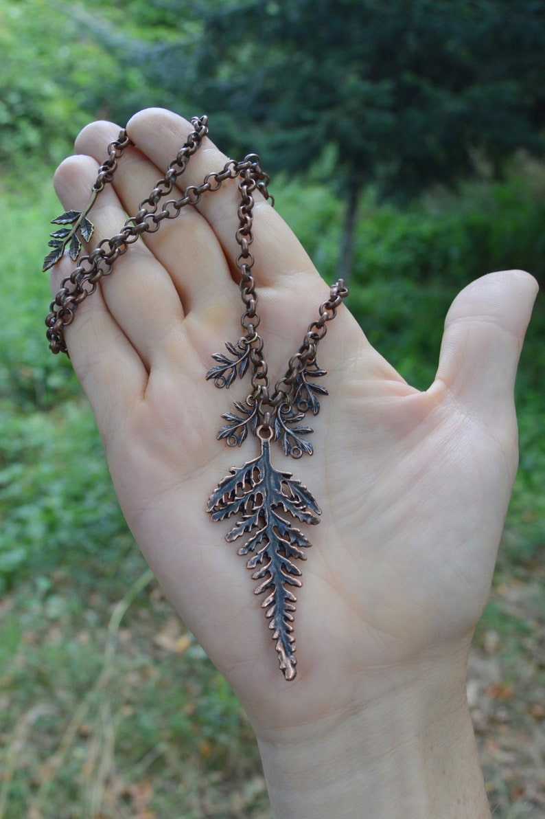 Copper leaf pendant displayed on a hand. Large central fern leaf is 2 1/2 inches long, with smaller ferns either side hanging from a thick copper chain. Behind is lush green of a summer forest.