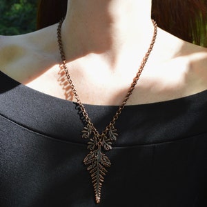 Copper leaf necklace made of copper with large fern leaf. Photo show it being worn against a black dress. The chain is long, thick and made of round links, and hangs in a low V shape. 4 tiny fern leaves hang either side of the large leaf.