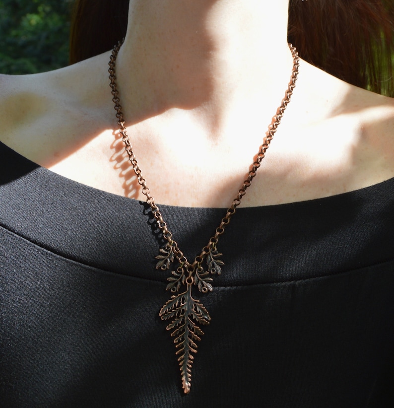 Copper leaf necklace made of copper with large fern leaf. Photo show it being worn against a black dress. The chain is long, thick and made of round links, and hangs in a low V shape. 4 tiny fern leaves hang either side of the large leaf.