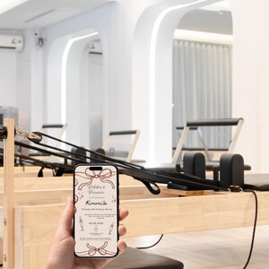May include: A smartphone displays a Pilates birthday party invitation with a bow design. The phone is held in front of Pilates equipment in a bright, modern studio. The invitation text reads "Pilates & Prosecco" and "Kimmie's Pilates Birthday Party".