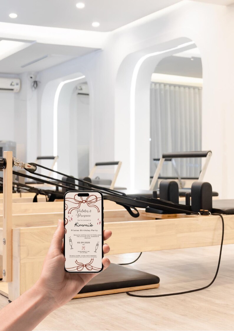May include: A smartphone displays a Pilates birthday party invitation with a bow design. The phone is held in front of Pilates equipment in a bright, modern studio. The invitation text reads "Pilates & Prosecco" and "Kimmie's Pilates Birthday Party".