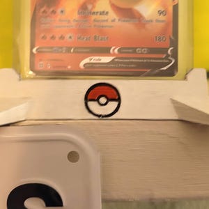 May include: White card display stand with a red and black Pokemon ball logo. The stand is holding a Pokemon trading card. A Texas license plate is visible in the foreground.